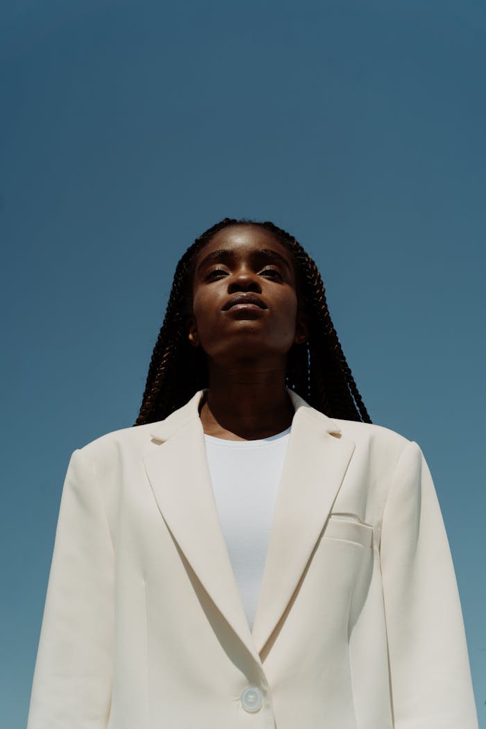 Low angle portrait of a confident young woman in a white blazer against a clear blue sky.
